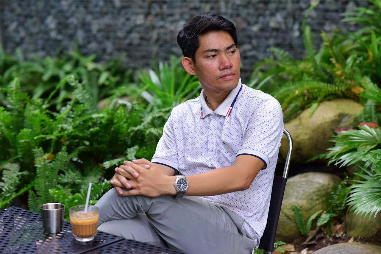 who-we-are A man in casual attire sitting outdoors in a garden, enjoying a beverage.